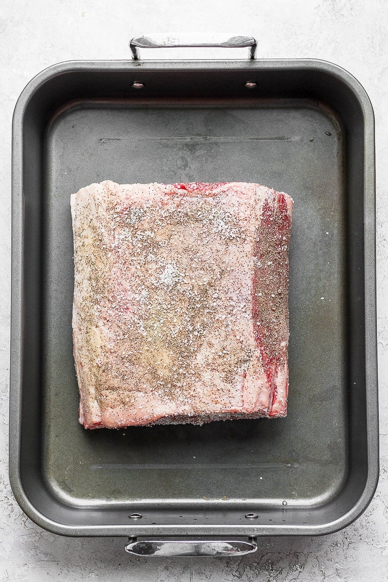 Prime rib with salt and pepper rub sitting in roasting pan, uncooked. 