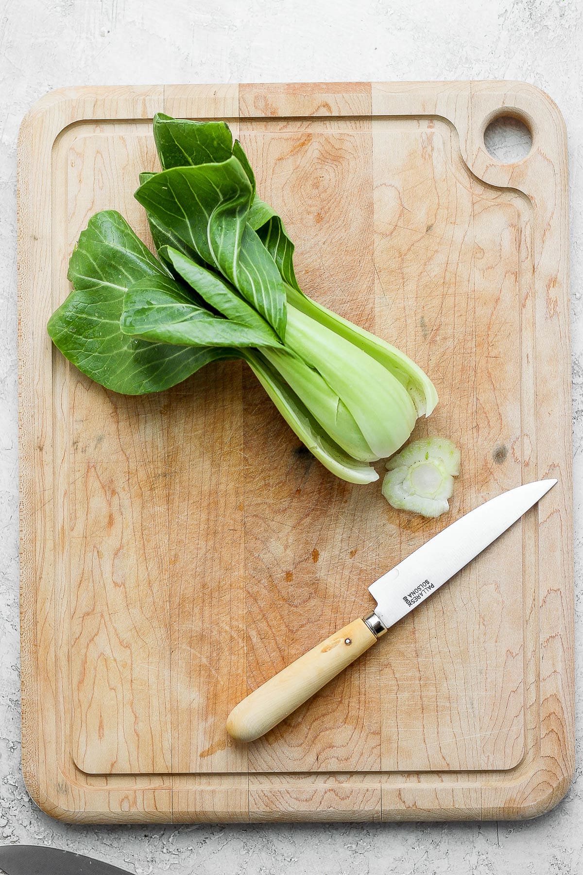 A large wooden cutting board with one baby bok choy on top, a wooden knife and the end of the bok choy cut off. 