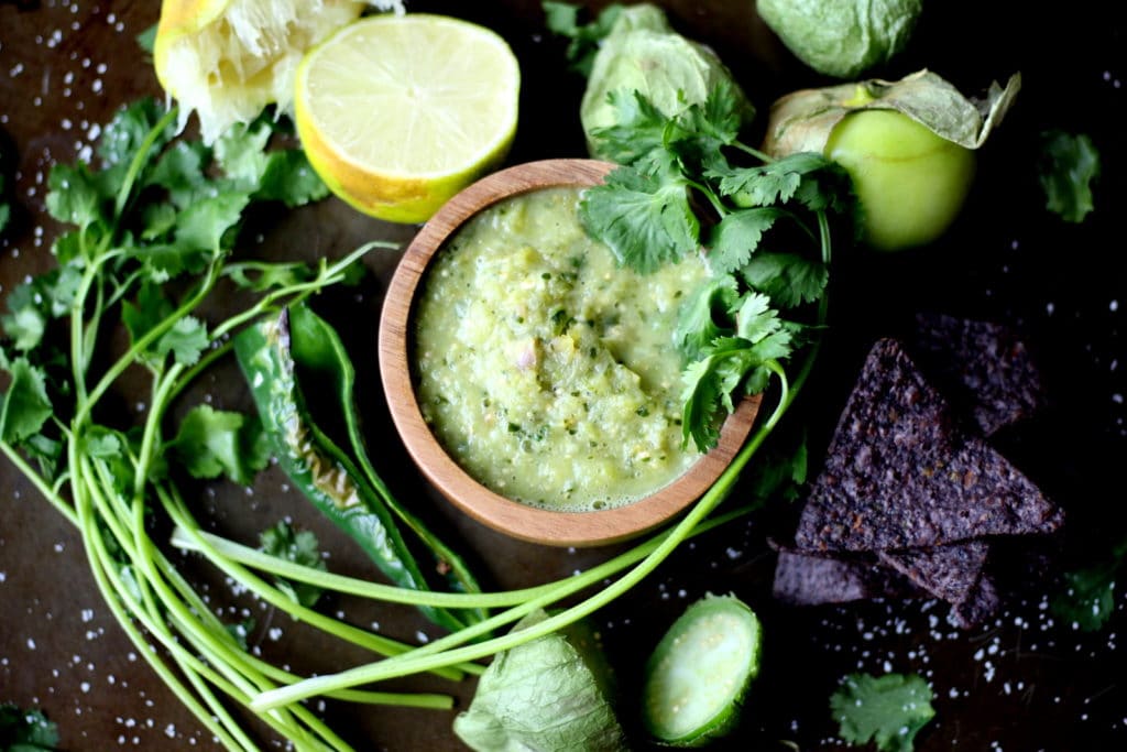 Small wooden bowl of salsa verde surrounded by blue corn chips, tomatillos, limes and fresh cilantro. 