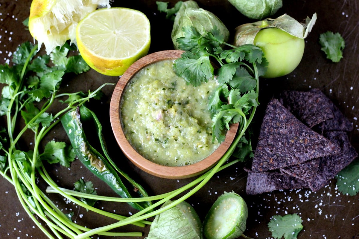 Small wooden bowl of salsa verde surrounded by blue corn chips, tomatillos, limes and fresh cilantro. 