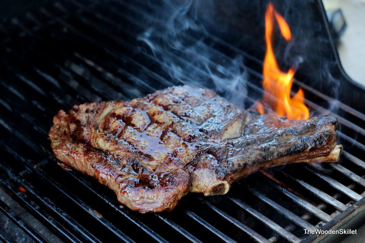 A bone-in ribeye steak on the grill with a flame behind it. 
