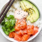 A salmon sushi bowl with salmon, cilantro, avocado, spicy mayo and sesame seeds with two chopsticks resting on the side of the bowl.