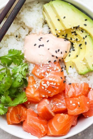 A salmon sushi bowl with salmon, cilantro, avocado, spicy mayo and sesame seeds with two chopsticks resting on the side of the bowl.