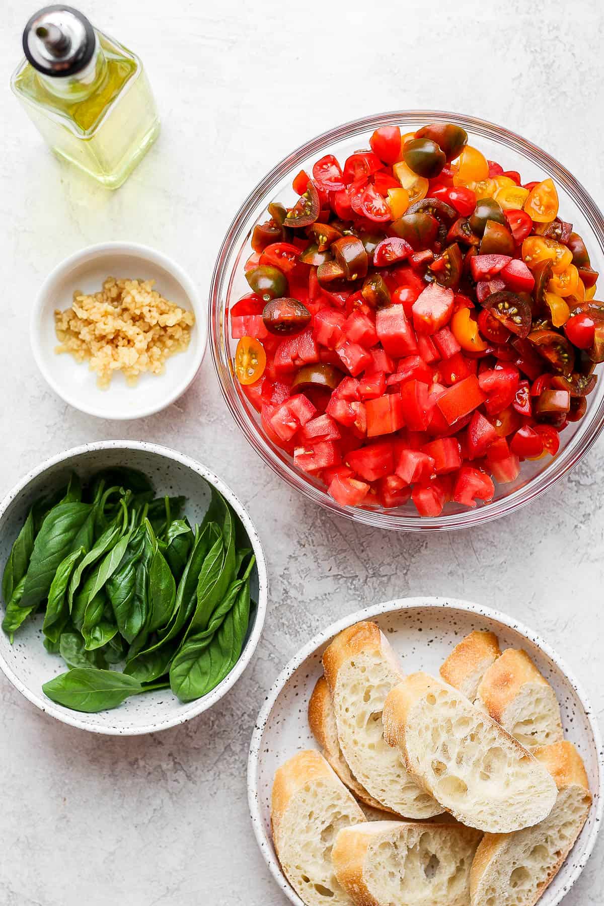 Bowls of cut tomatoes, garlic, basil leaves, and baguette slices on a plate.