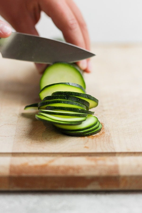 One zucchini being cut into thin slices on a cutting board.