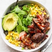 Pulled pork with a drizzling of bbq sauce in a bowl with lettuce, corn kernels, half of a sliced avocado and a fork sticking out the side.