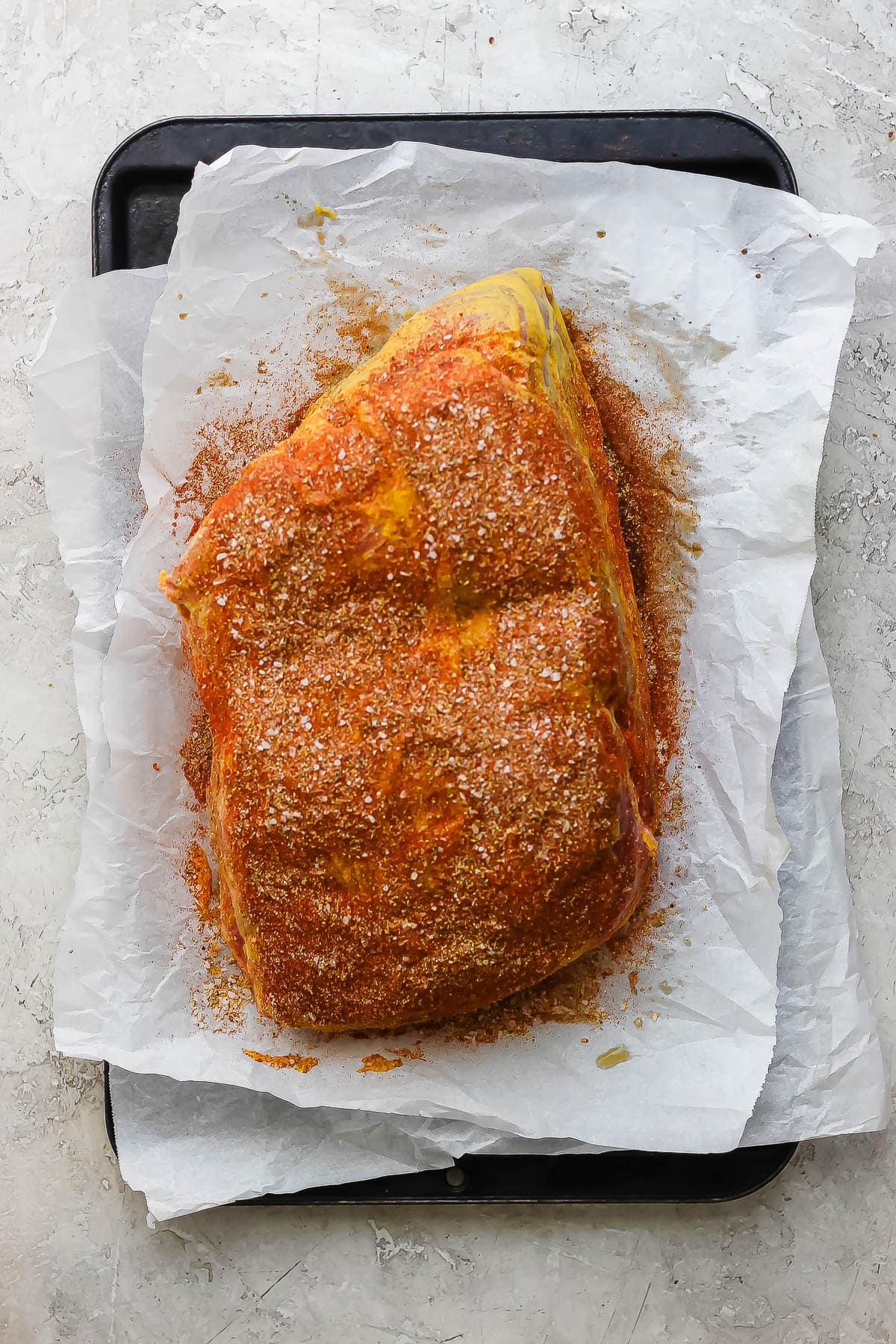 A pork shoulder in a baking sheet covered yellow mustard and then the dry rub on top.