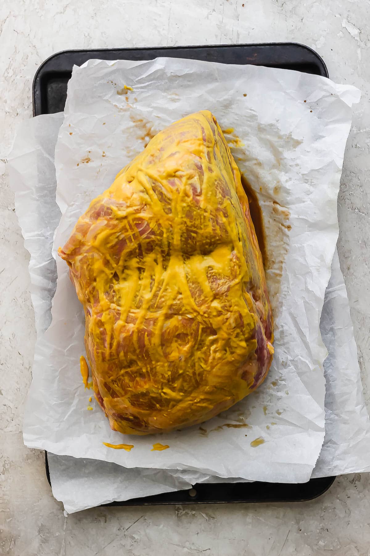 A pork shoulder on a baking sheet covered in yellow mustard.