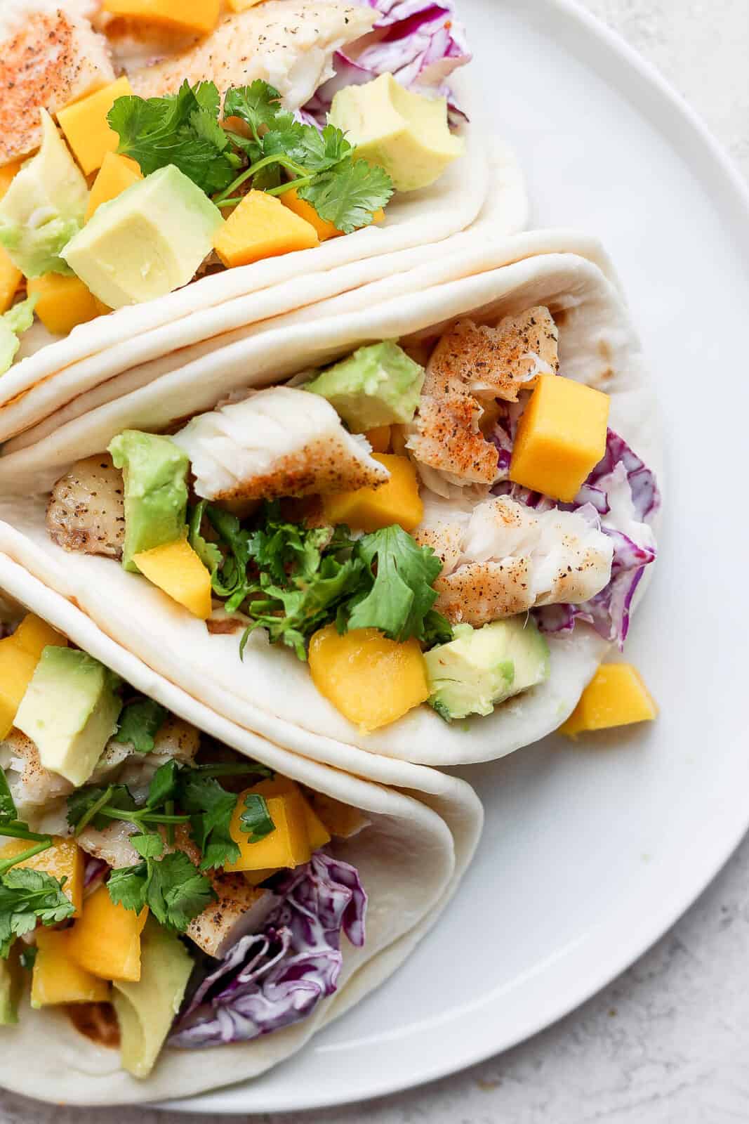 Three tilapia fish tacos with mango and avocado on a white plate. 