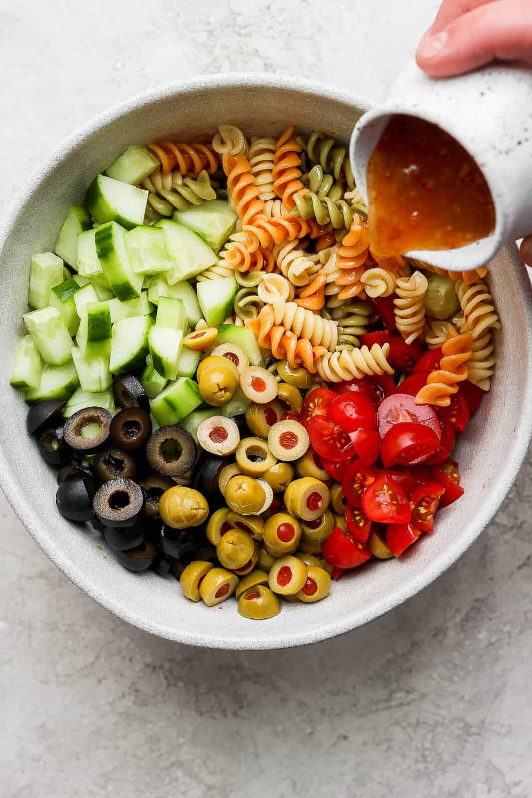Cooked noodles in a bowl with pasta salad ingredients and someone pouring dressing on top. 