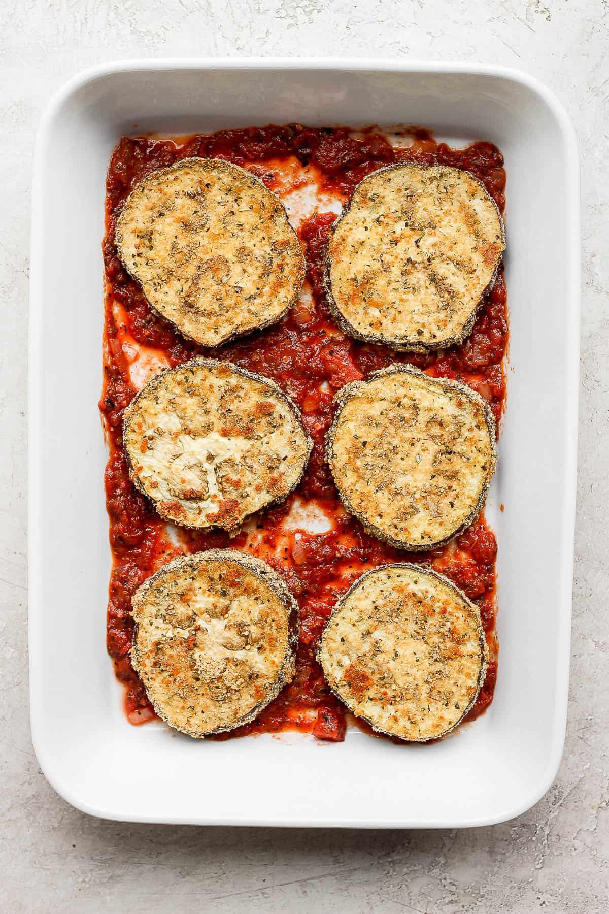 A large baking dish with pasta sauce in the bottom and six breaded eggplant slices on top. 