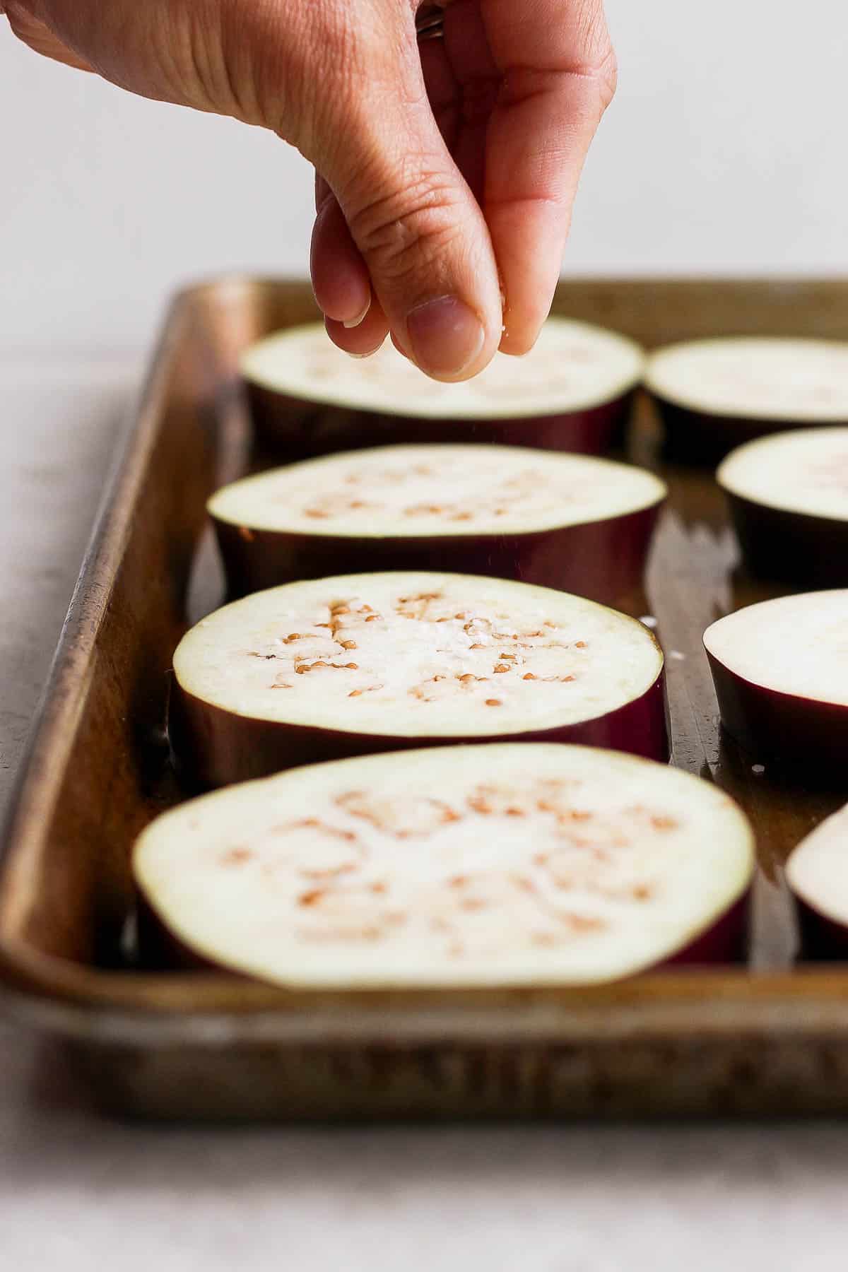 Sliced eggplant on a baking sheet with someone sprinkling salt on top of each. 