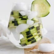 A cocktail glass with a cucumber vodka and water over ice and garnished with fresh cucumber slices.