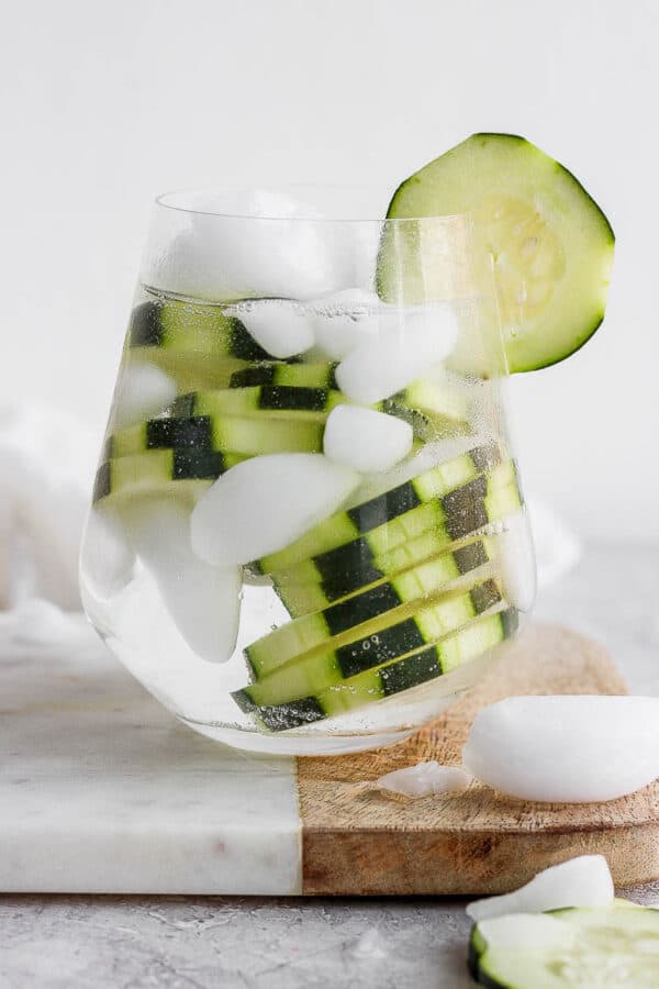 A cocktail glass with a cucumber vodka and water over ice and garnished with fresh cucumber slices.