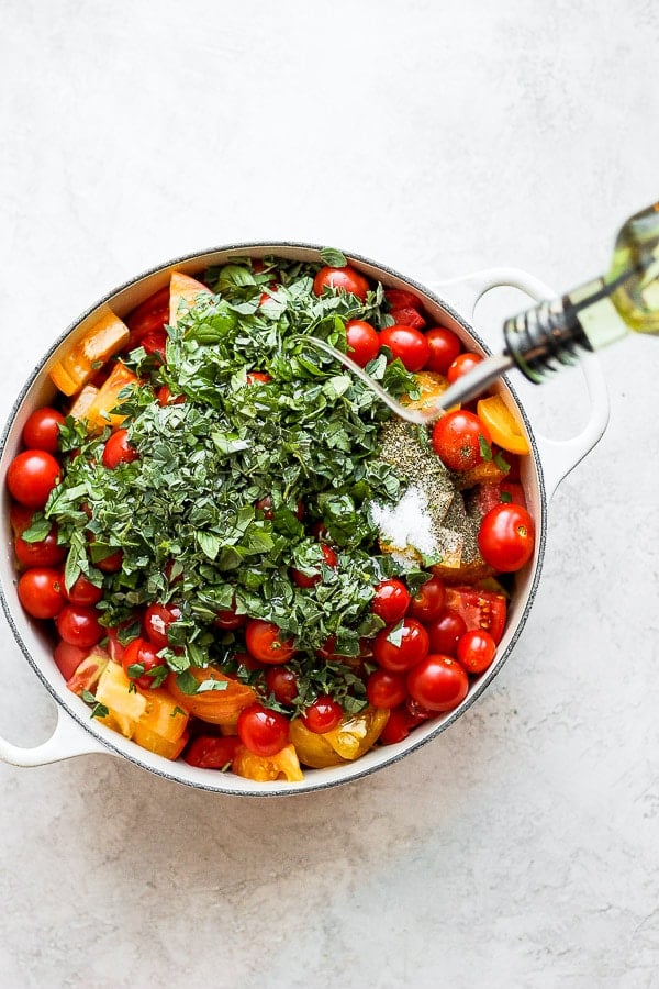 Someone pouring olive oil into dutch oven filled with tomatoes and herbs. 