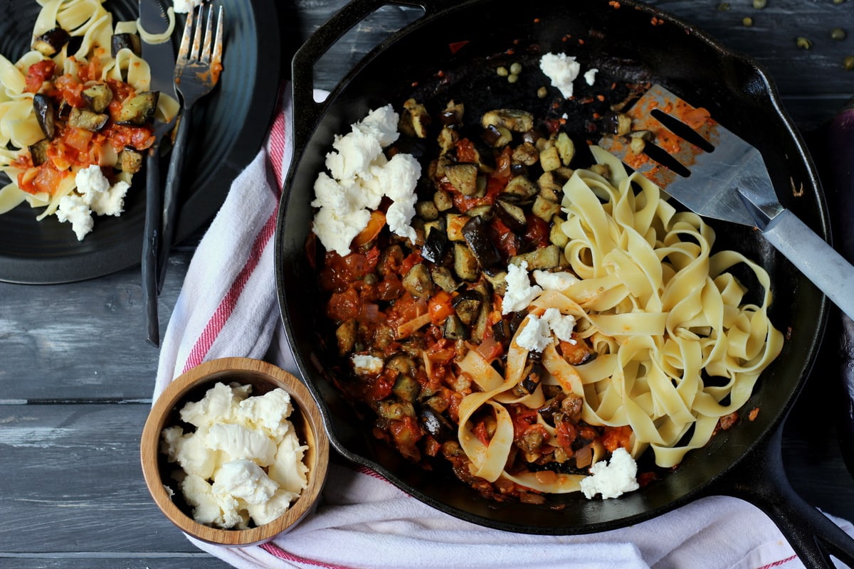 Eggplant ragu in a cast iron skillet with noodles being added.