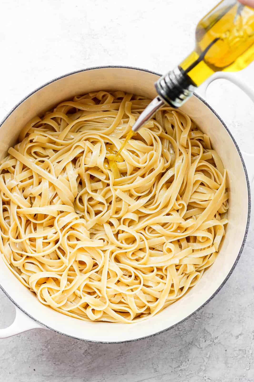 Someone drizzling olive oil into a pan of cooked noodles. 