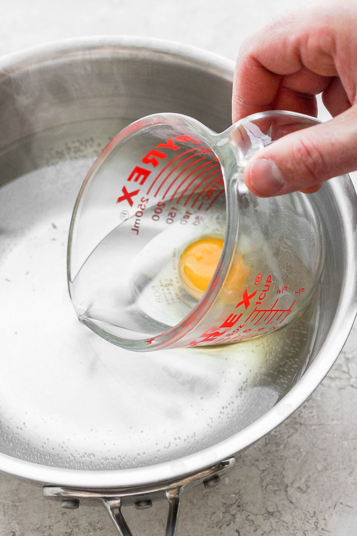 An egg in a glass measuring cup being lowered into simmering water.