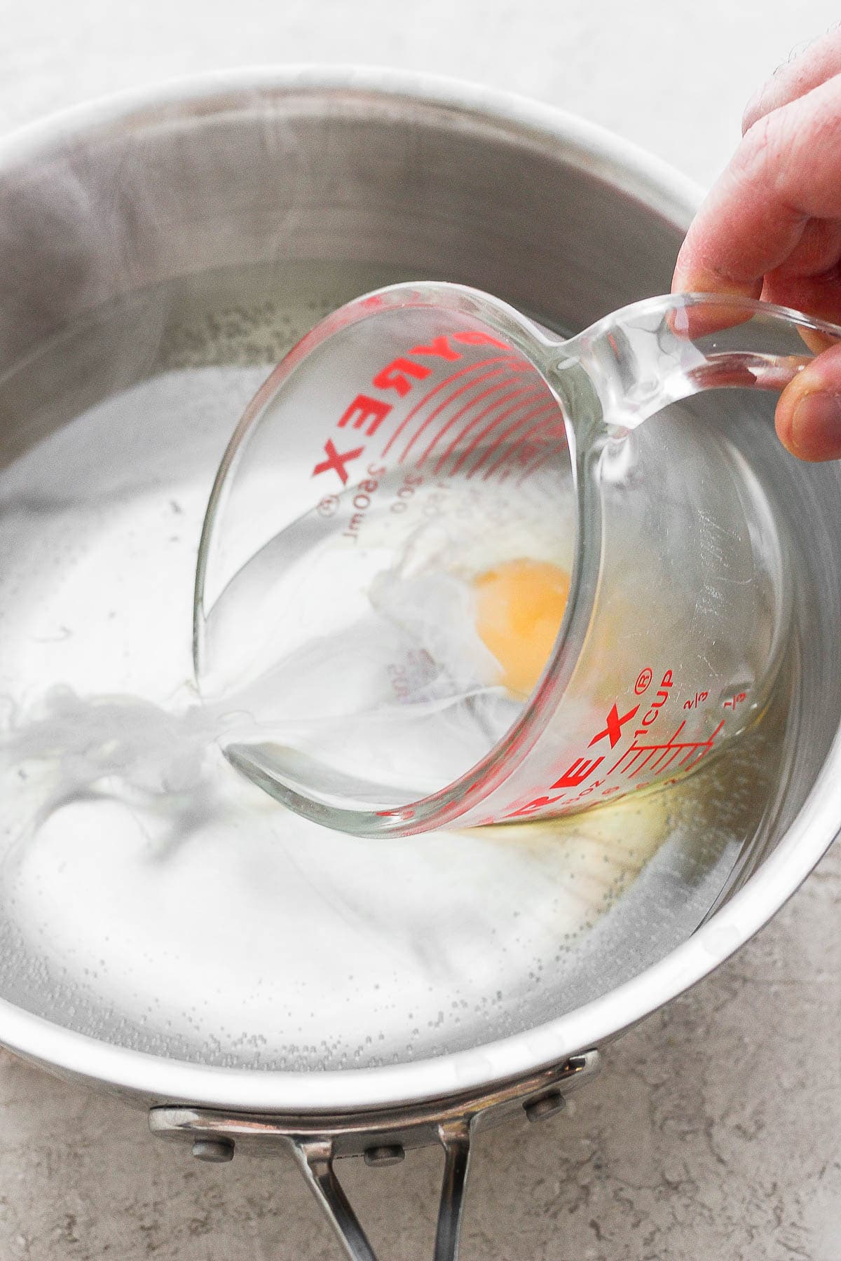 A hand slowly pouring a raw egg into a saucepan with hot water in it 