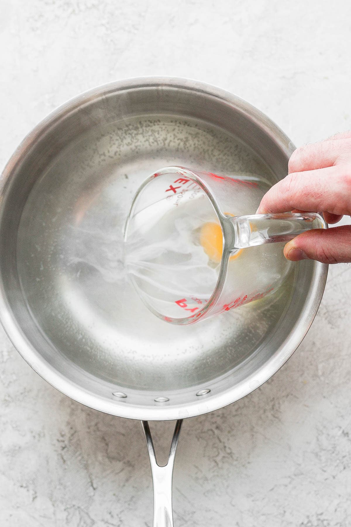 A hand pouring a raw egg into a saucepan with hot water in it. 