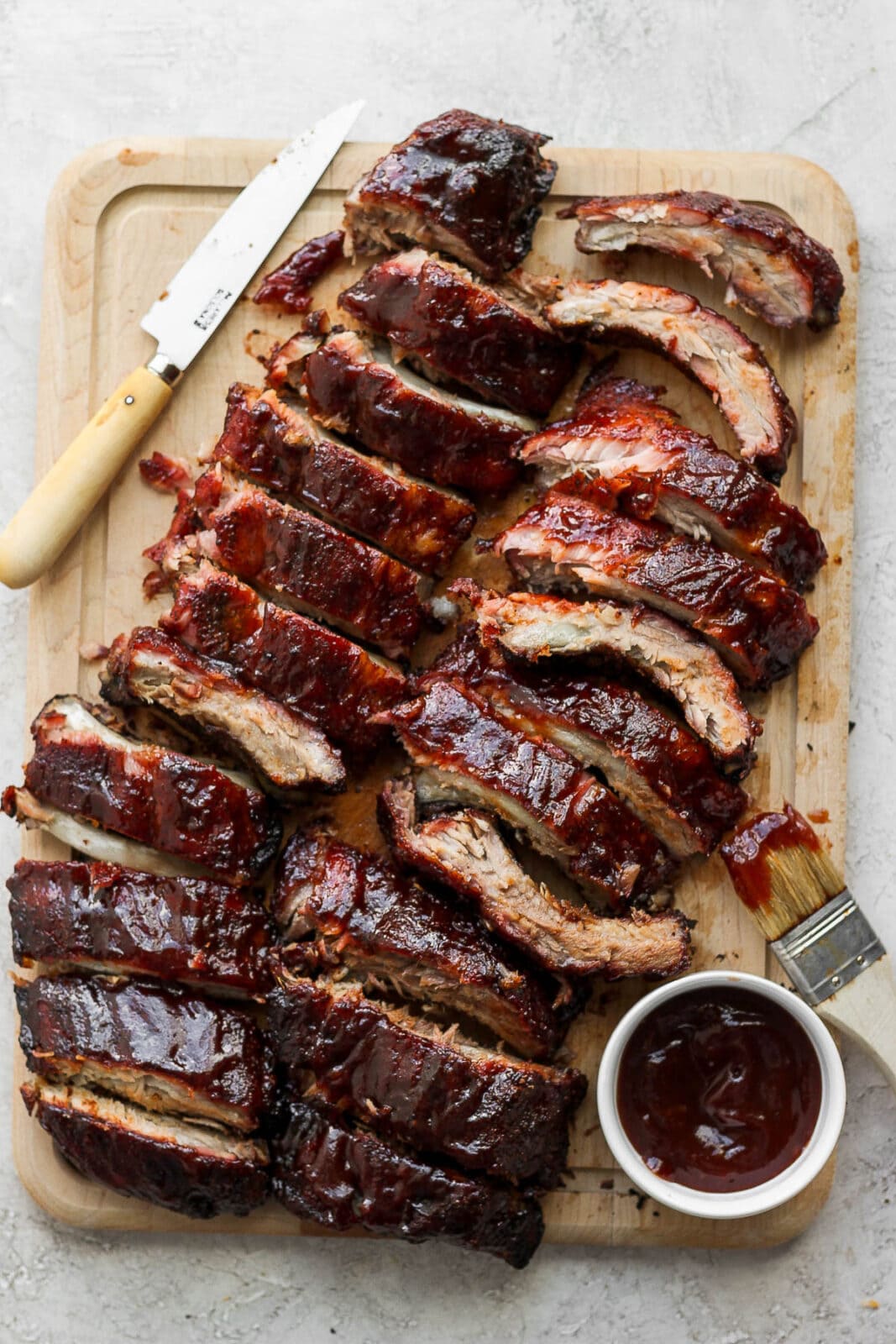 A wooden cutting board with two racks of ribs on top of it cut up and a wooden knife next to them. 