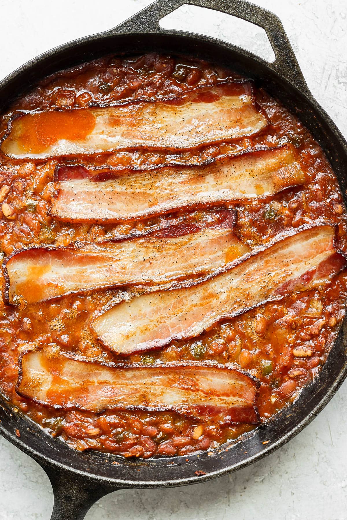 Cast iron skillet filled with smoked baked beans with bacon laying across the top. 
