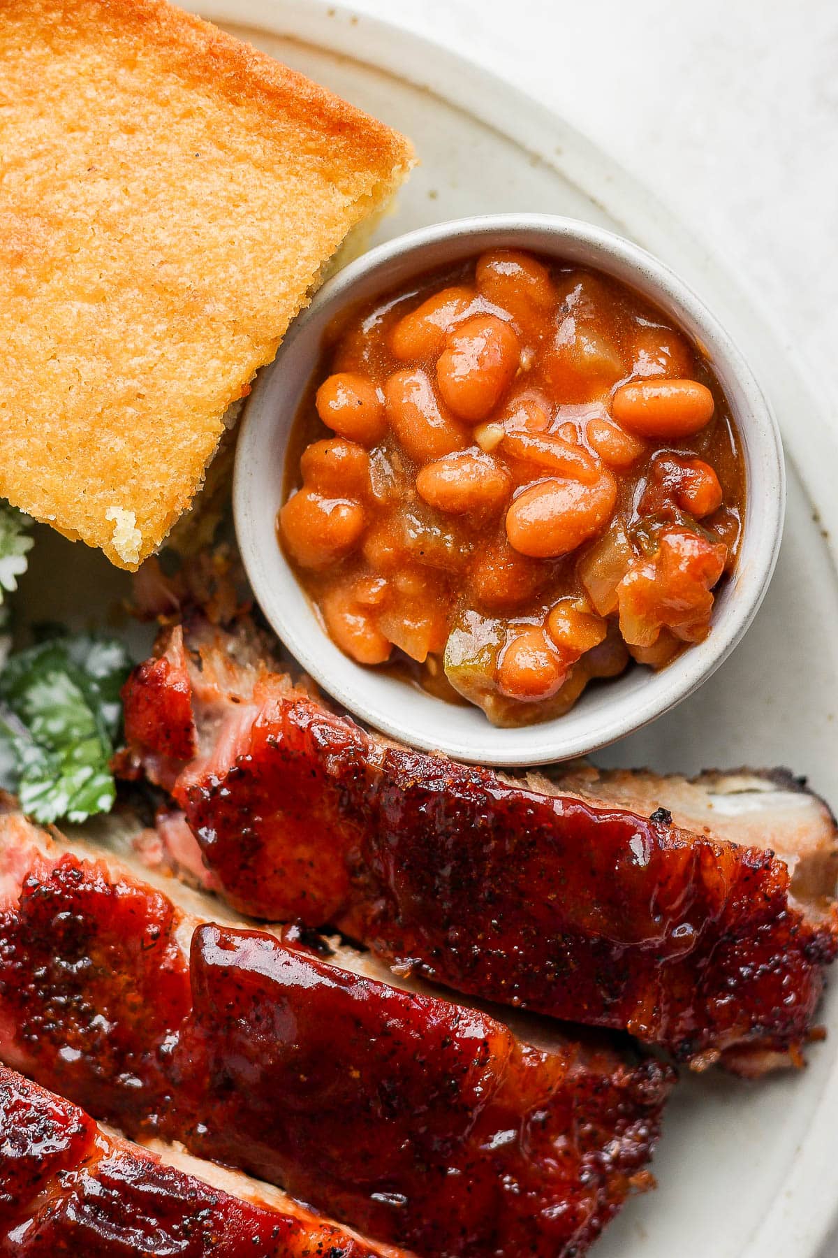 Smoked Baked Beans in a small bowl sitting on top of plate next to smoked rib and cornbread. 