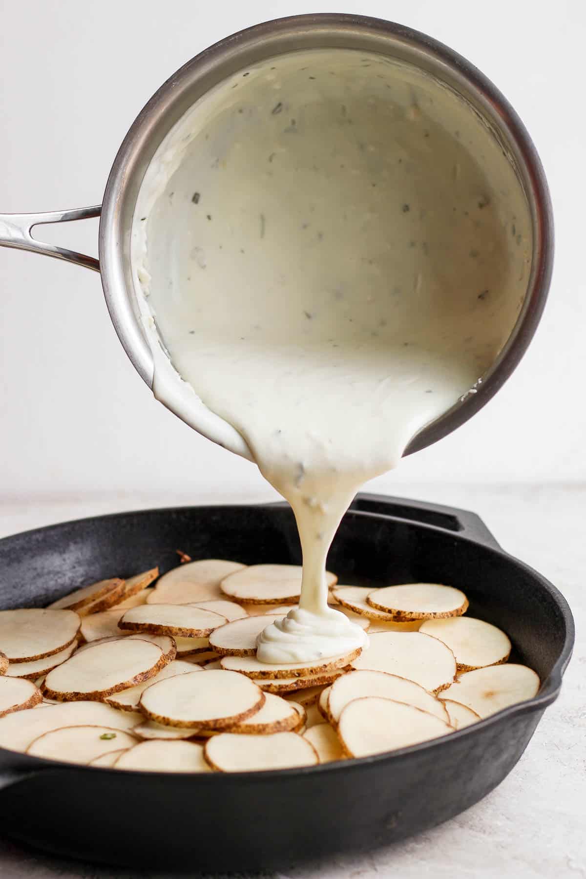 The cheese sauce being poured over the sliced potatoes in a cast iron skillet.