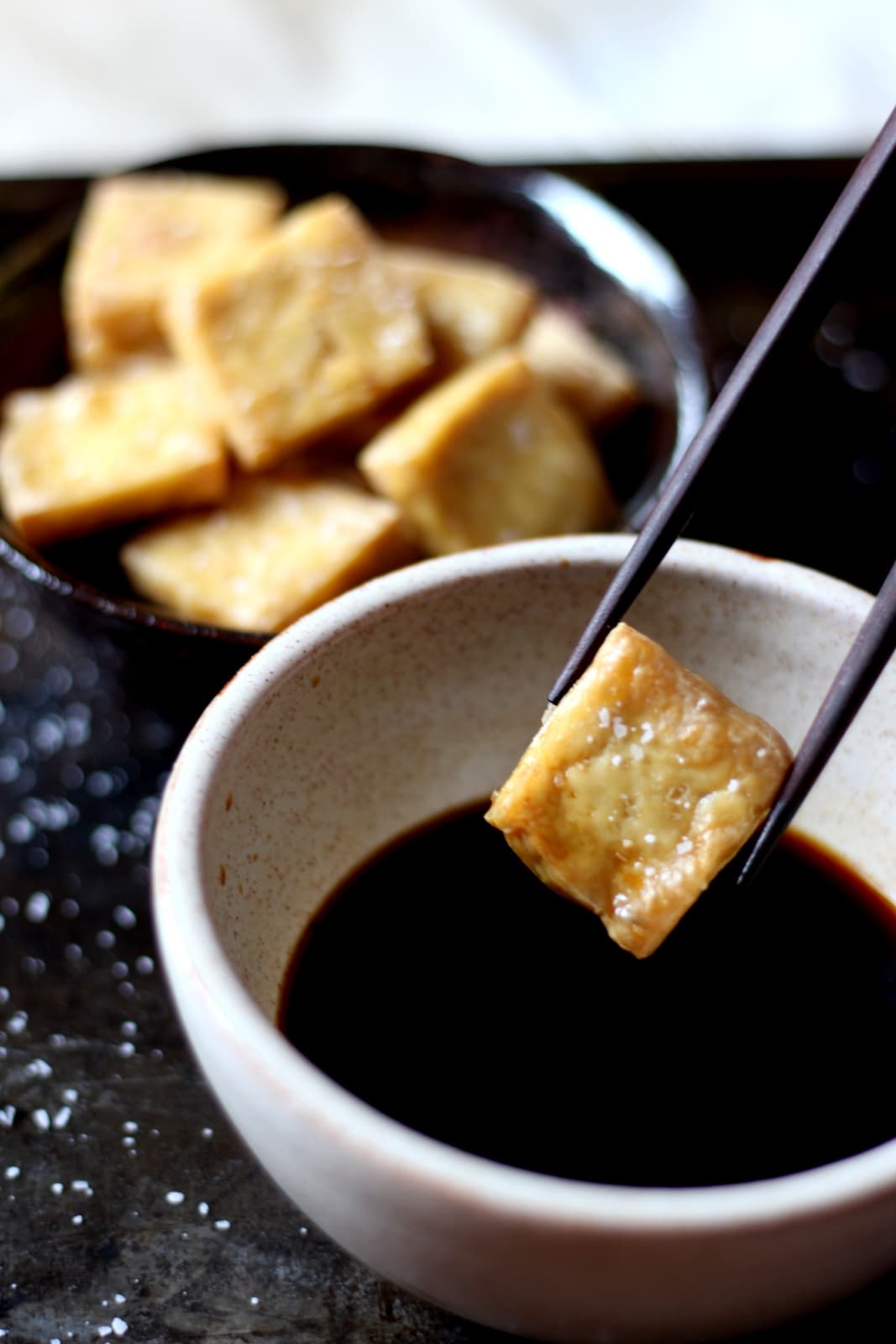 A pair of chopsticks holding a piece of cooked tofu over a bowl of soy sauce. 