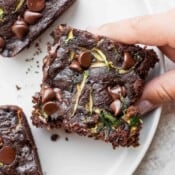 Double chocolate zucchini brownies on a white plate with a hand grabbing one of them.