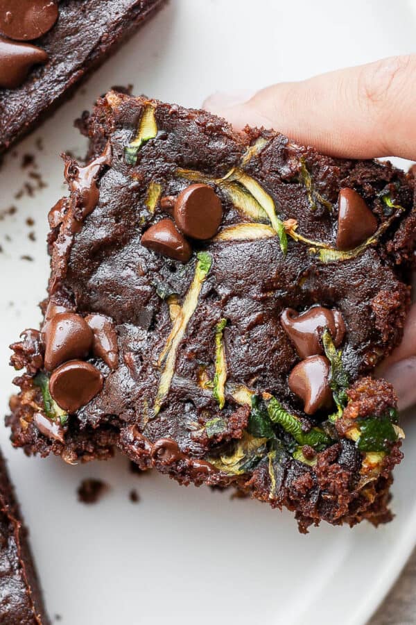 Double chocolate zucchini brownies on a white plate with a hand grabbing one of them.