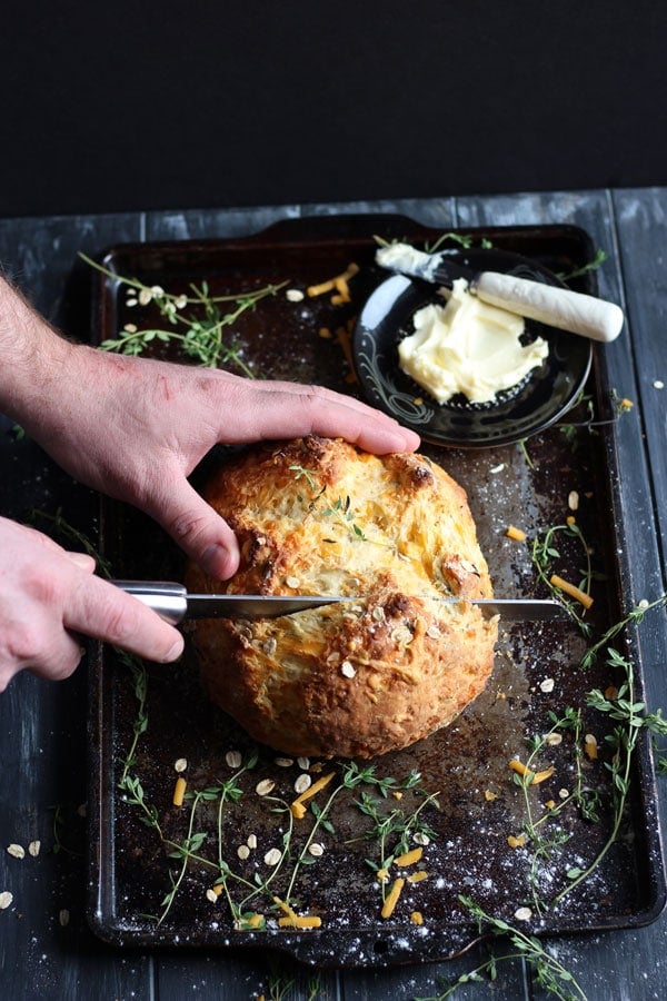 Cheesy Irish Soda Bread + Fresh Thyme and Rosemary. A cheesy twist on the classic Irish tradition - the perfect recipe for St. Patrick's Day! thewoodenskillet.com #stpatricksday #irishsodabread The Irish soda bread being cut with a bread knife.
