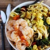 A bowl with fettuccine, shrimp, kale and roasted turmeric cauliflower on a brown board with silverware.