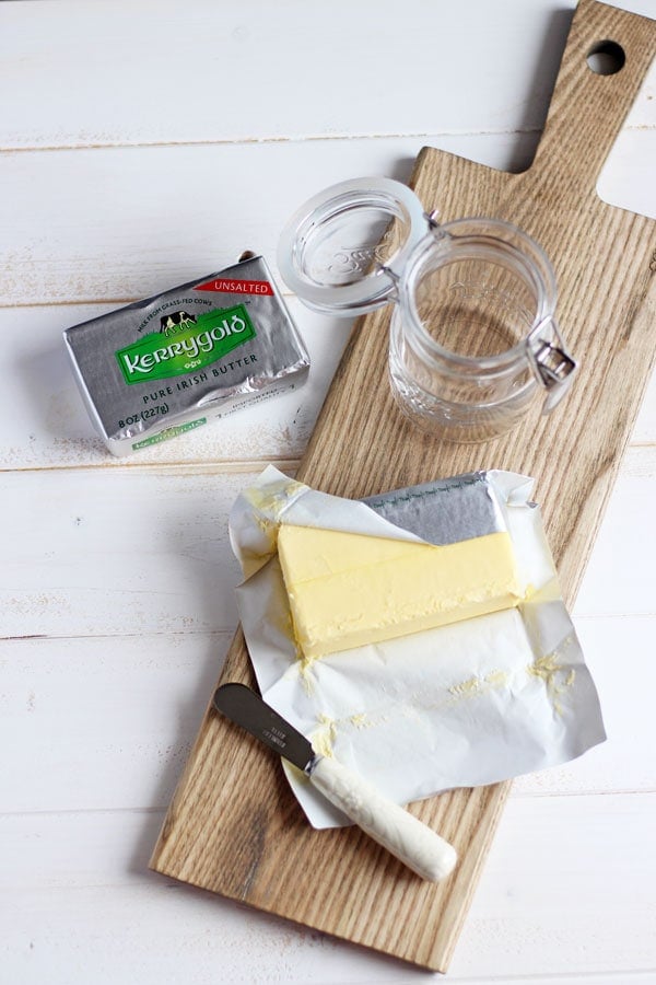 A wooden board set diagonally with an empty jar for ghee, an unopened Kerrygold bar of butter and an opened Kerrygold butter.