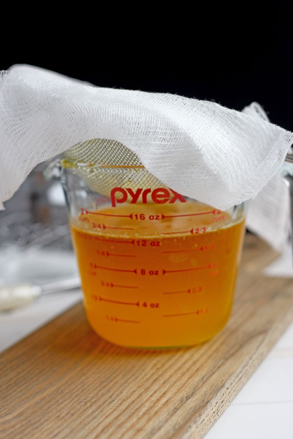A glass measuring cup filled with melted ghee with a metal sieve and cheesecloth on top for straining.