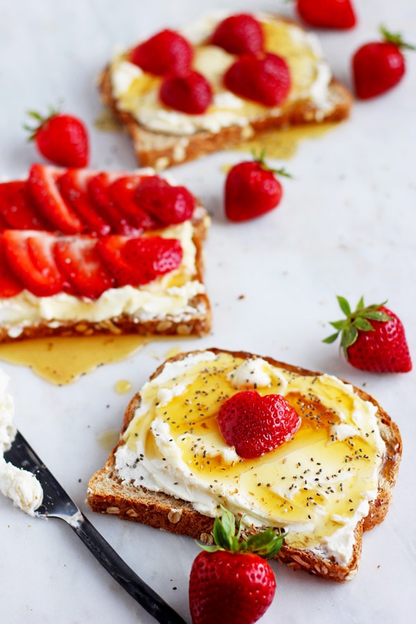 A piece of toast with whipped cream cheese, chia seeds and strawberries on a marble board with strawberries scattered around. 
