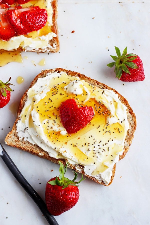 A piece of toast with whipped cream cheese, chia seeds and strawberries on a marble board with strawberries scattered around. 