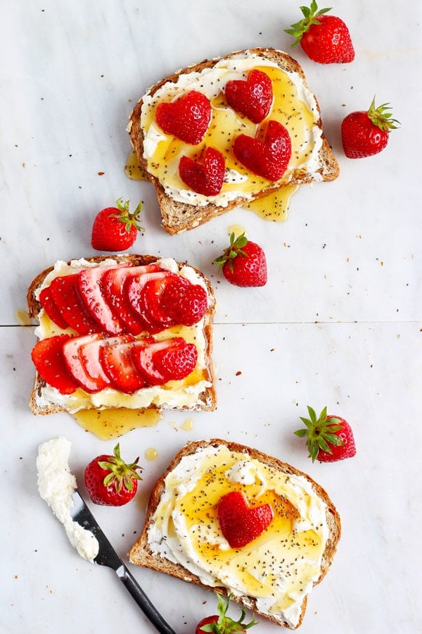 Three pieces of toast with whipped cream cheese, chia seeds and strawberries on a marble board with strawberries scattered around. 