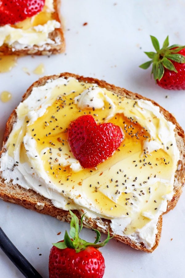 A piece of toast with whipped cream cheese, chia seeds and strawberries on a marble board with strawberries scattered around. 