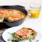 A slice of a sausage and mushroom frittata on a small white plate with the whole frittata and glass of orange juice in the background.