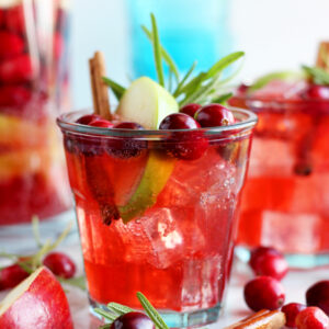 A glass of cranberry cinnamon apple kombucha mocktail with a stick of cinnamon, apple slice, and rosemary for garnish.