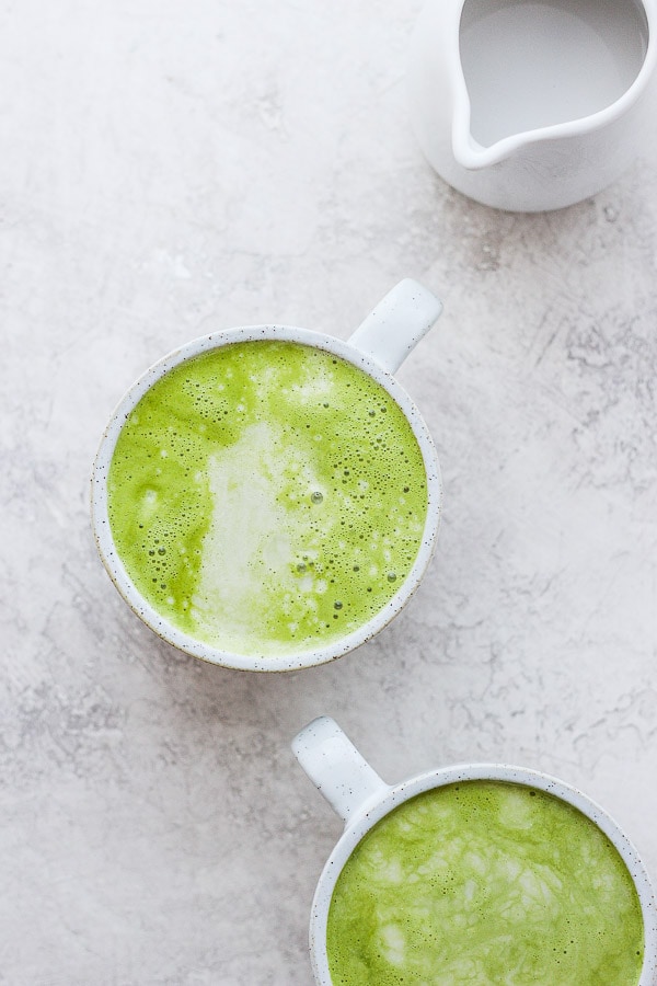 Two mugs of matcha lattes and a small pitcher of almond milk. 