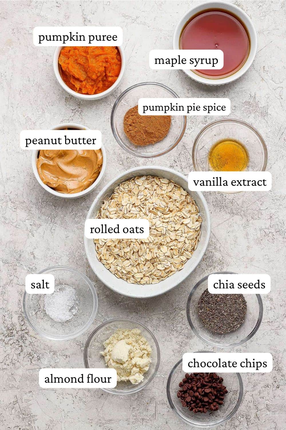 Pumpkin energy ball ingredients in individual bowls; pumpkin puree, maple syrup, pumpkin pie spice, peanut butter, vanilla extract, rolled oats, salt, almond flour, chia seeds, and mini chocolate chips.