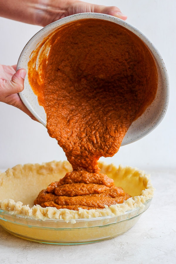 Two hands holding a bowl pouring pumpkin pie puree into a pie crust. 