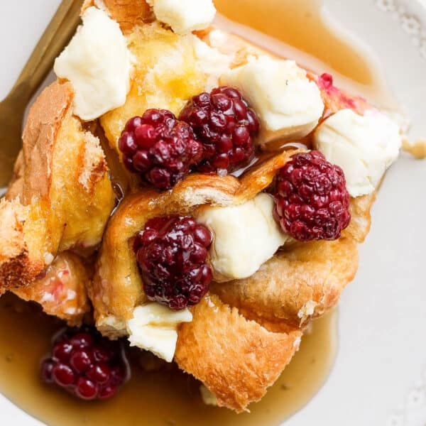 Top shot of a piece of french toast bake with chunks of cream cheese and blackberries on top and syrup on the plate.
