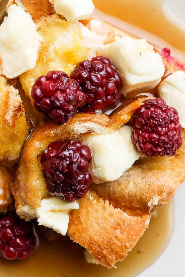 Top shot of a piece of french toast bake with chunks of cream cheese and blackberries on top and syrup on the plate.