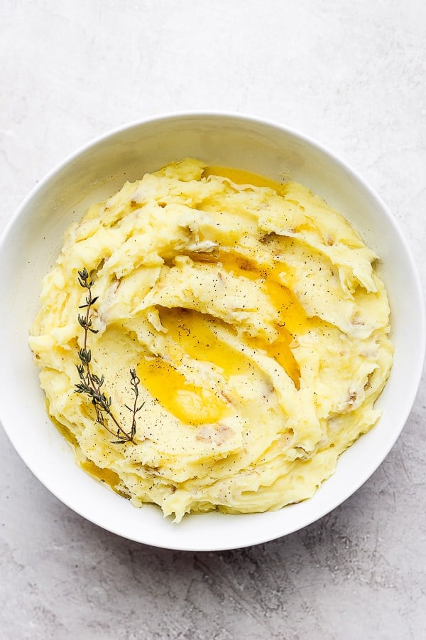 A white bowl of creamy mashed potatoes with a bit of fresh thyme on top.