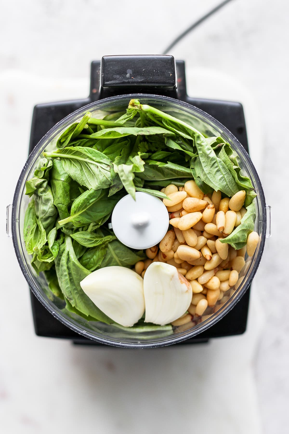 A top shot of a food processor filled with fresh basil, pine nuts and garlic cloves.