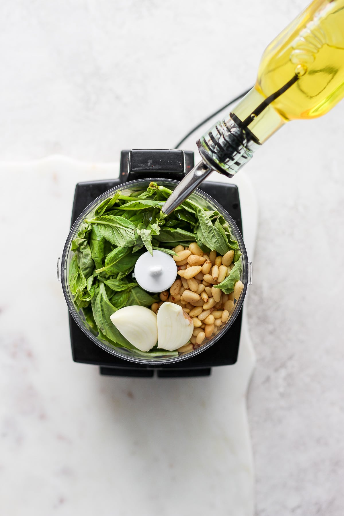 Top shot of a food processor filled with fresh basil, pine nuts, and garlic with someone drizzling olive oil into it. 