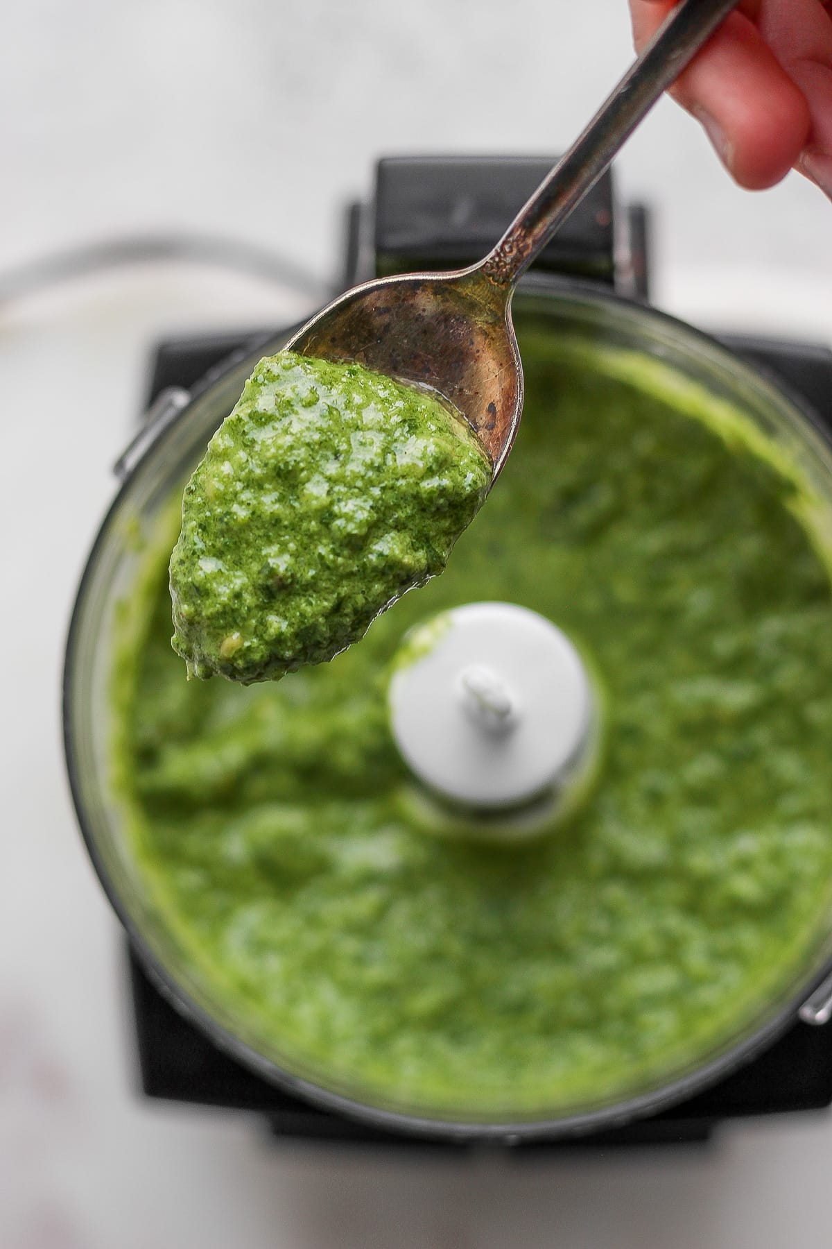 In the background a food processor filled with vegan pesto and in the foreground a spoon of pesto. 